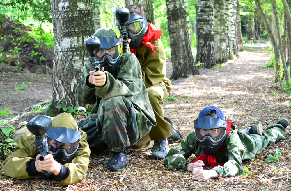 Мальчики в лесу в камуфляжных костюмах и защитных масках. Целятся из пейнтобольных ружей в камеру